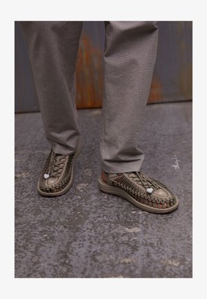 Sandali intrecciati beige con suole spesse, caratterizzati da un intricato motivo in pizzo e dettagli accentuati in bianco, abbinati a pantaloni a quadri grigi.