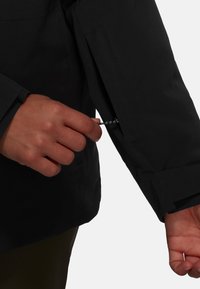 Schwarze wasserdichte Jacke mit glatter Textur, ausgestattet mit einer versteckten Reißverschlusstasche am Ärmel und einem engen Bunddesign.