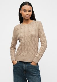 Pull en tricot beige avec un motif en câble, col rond, poignets et ourlet côtelés. Porté avec un jean en denim bleu, les mains dans les poches.