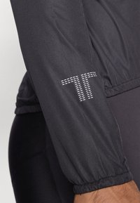 Crna atletska jakna s teksturiranim, elastičnim rubom i reflektirajućim logotipom "TT". Materijal izgleda glatko s mat finišem.