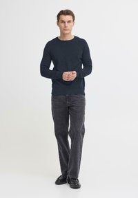 Marineblauwe gebreide trui met lange mouwen, ronde hals en geribde zoom, gecombineerd met grijze jeans met rechte pijpen en zwarte schoenen. Minimalistische stijl.