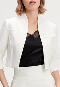 Veste blanche cropped avec un col haut et des manches trois-quarts, portée sur un caraco noir en satin avec des garnitures en dentelle.