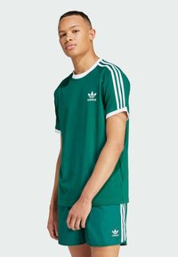 Green cotton t-shirt with white ribbed crew neck and sleeve cuffs, featuring the Adidas logo and three white stripes on the shoulders.
