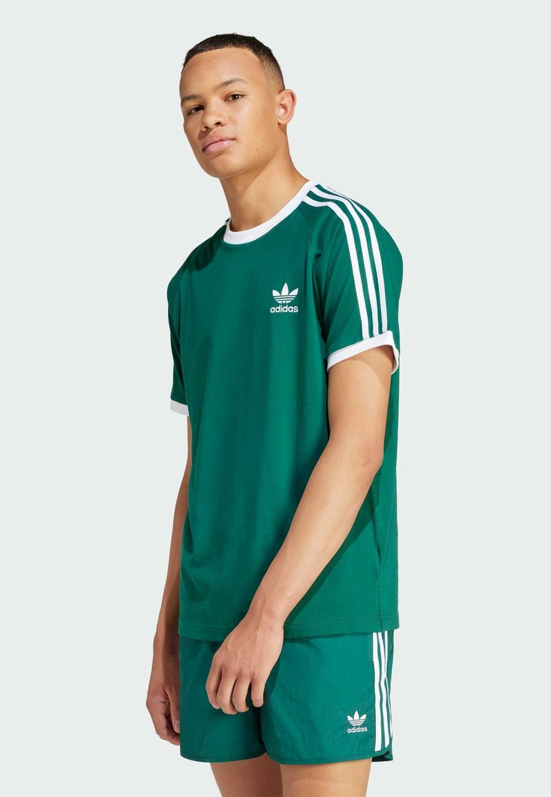 Green cotton t-shirt with white ribbed crew neck and sleeve cuffs, featuring the Adidas logo and three white stripes on the shoulders.