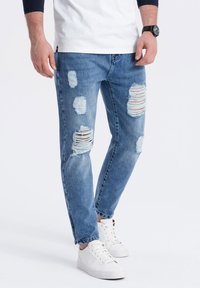 Jeans rotos de color azul claro con múltiples parches desgastados y un corte recto, combinados con zapatillas blancas y una camisa blanca y azul marino.