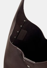 Sac fourre-tout en cuir marron avec une texture douce, présentant des accents en métal et une étiquette à l'intérieur. Design à ouvert sur le dessus avec des bords lisses.