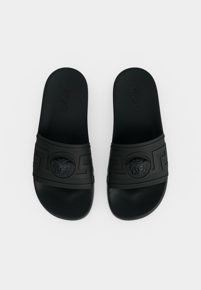 Pair of black slide sandals with embossed Medusa head and Greek key design on the straps, viewed from above.