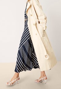 Gabardina beige, vestido azul marino con rayas diagonales blancas y sandalias blancas de tacón con tiras. Telas suaves, formas fluidas, herrajes mínimos.