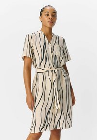 Beige jurk met korte mouwen, voorzien van zwarte golvende lijnen, knoopsluiting aan de voorkant en een tailleband. Heeft een borstzak en een ontspannen pasvorm.