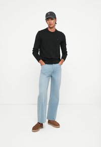Suéter de punto negro, jeans rectos de color azul claro, mocasines marrones y una gorra oscura con logotipo. Vestimenta simple y casual.