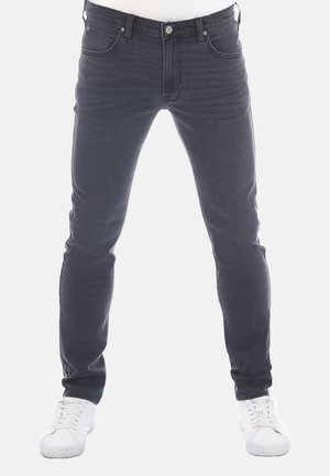 Homme portant un jean slim foncé et des baskets blanches, debout les jambes légèrement écartées sur un fond uni.