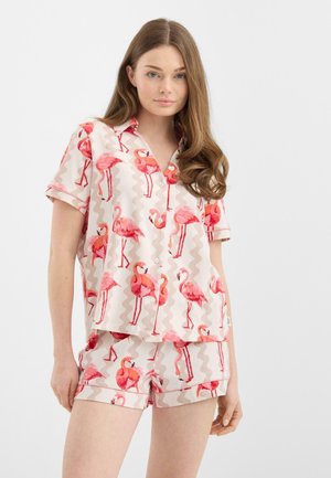 Vrouw draagt een beige shirt met korte mouwen en bijpassende korte broek met roze flamingoprints, staand tegen een effen witte achtergrond.