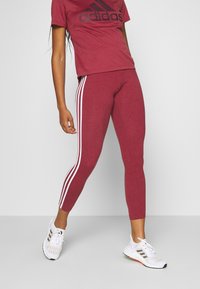 Maroon urheiluleggingsit valkoisilla sivuviivoilla, yhdessä maroon t-paidan kanssa, jossa on musta logo, ja valkoiset lenkkarit. Sileä tekstuuri.