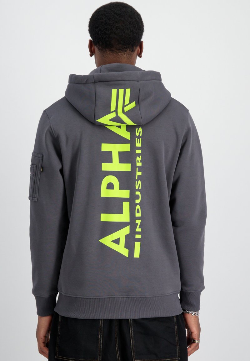 Felpa grigia con un vistoso logo giallo neon "ALPHA INDUSTRIES" sul retro, cappuccio regolabile e polsini a coste. Una tasca con zip su un lato.
