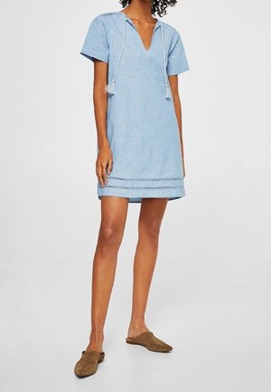 Femme portant une robe bleu clair à manches courtes avec des liens à pompons et des chaussures marron sans lacets, debout devant un fond uni.