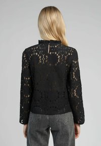 Top en dentelle noire à manches longues, motif floral, col froncé, fermeture à boutons au dos, porté avec un jean taille haute gris.