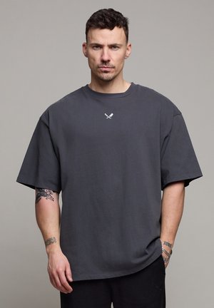 Man met kort haar die een oversized donkergrijs T-shirt draagt met een klein ontwerp van gekruiste messen, tatoeages op zijn armen, staand tegen een effen achtergrond.