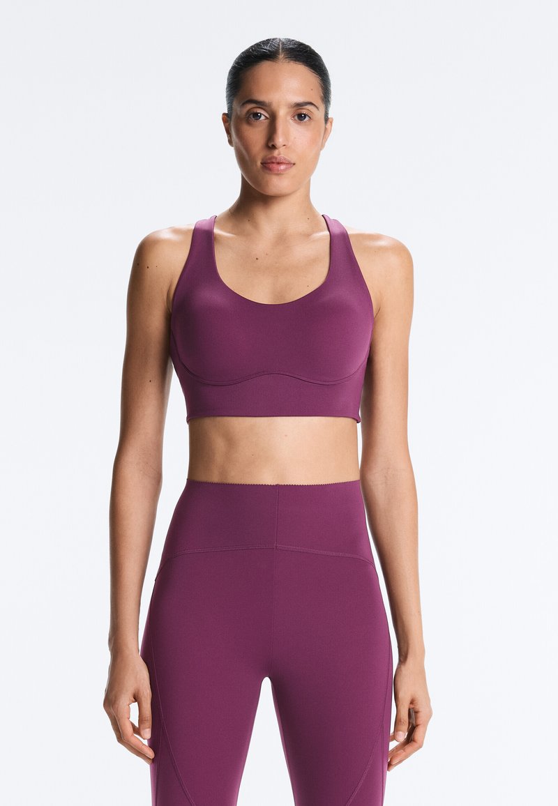 Kvinde iført en matchende lilla sports-bh og højtaljede leggings, stående oprejst mod en ensartet lys baggrund.