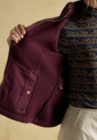 Burgunderfarbene Zip-Jacke mit weichem Fleece-Innenfutter, ausgestattet mit einer Seitentasche und goldenen Beschlägen. Über einem blau gemusterten Oberteil getragen.