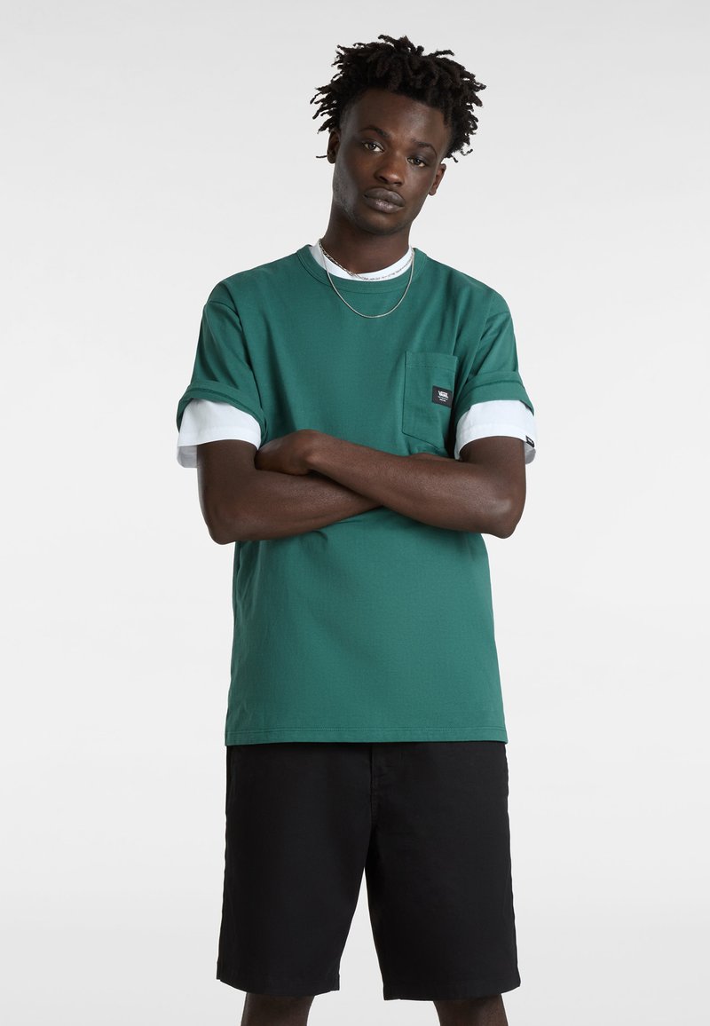 Camiseta verde de manga corta con puños blancos enrollados y un bolsillo frontal, combinada con shorts negros. Presenta una etiqueta de la marca en el bolsillo.