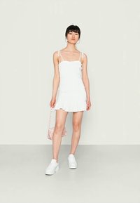 Vestido mini branco com bainha franzida, alças finas e decote com babados. Modelo usa ténis brancos e segura uma bolsa floral.