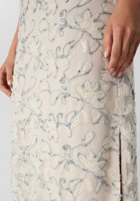Tissu beige avec des motifs floraux brodés en bleu clair et des sequins, montrant une main reposant doucement sur le matériau près d'une fente sur le côté.