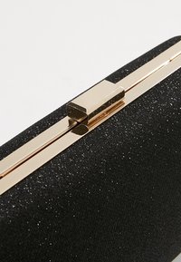 Pochette noire pailletée avec une fermeture à rabat rectangulaire brillante en or, présentée en gros plan.