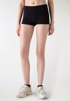 Persona che indossa pantaloncini aderenti neri e scarpe da ginnastica bianche, in piedi contro uno sfondo chiaro e uniforme.