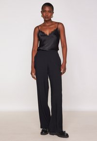 Vrouw in een zwarte satijnen cami met kantafwerking, hoge taille wijde zwarte broek, zwarte loafers en gouden hoepeloorbellen, staand tegen een effen achtergrond.