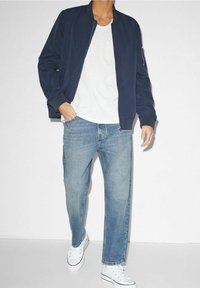 Marinblå bomberjacka med ribbad mudd, vit T-shirt under, ljusblå raka jeans och vita sneakers. Enkelt, avslappnat outfit.