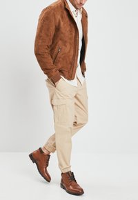 Giacca bomber in suede marrone, pantaloni cargo beige con polsini elasticizzati e stivali lacci marroni scuri, caratterizzati da materiali testurizzati e tasche utilitaristiche.