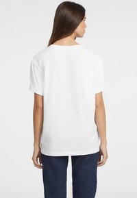 T-shirt bianca in cotone con maniche corte arrotolate, vestibilità ampia e scollo rotondo, vista da dietro con jeans blu scuro.