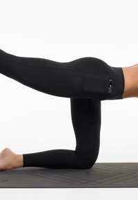 Leggings noirs taille haute avec une texture lisse et élastique. Présentent une poche latérale et un design ajusté, montrés dans une pose de yoga à genoux.