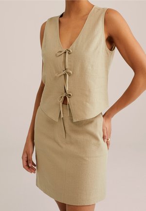 Femme portant un top en lin beige sans manches avec trois fermetures à nouer sur le devant et une jupe beige assortie avec une poche fendue à l'avant.