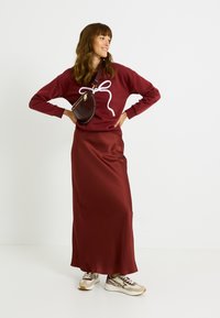 Sudadera burdeos con estampado de lazo blanco, falda maxi de satén burdeos y zapatillas beige. Bolso bandolera con tachuelas y detalle de cremallera.