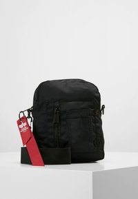 Črna torbica iz tkanine v obliki pravokotnika, z žepom z zadrgo spredaj, nastavljivim trakom in rdečo etiketo z blagovno znamko.