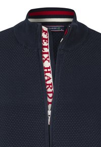 Mörkblå zip-up tröja med en texturerad mönstring, som har en hög krage och röda och vita randiga detaljer. Logotyp synlig på dragkedjan.