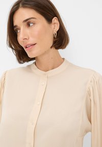 Beige blouse featuring a stand collar, button front, and gathered puff sleeves, made from a smooth, lightweight fabric.