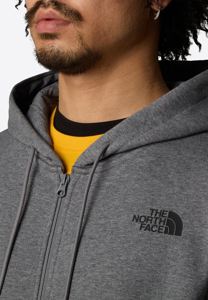 Sudadera gris con cremallera y cordones, superpuesta a una camiseta negra y amarilla. La textura parece suave y casual.