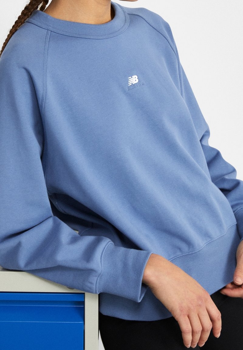 Personne portant un sweat-shirt bleu New Balance, appuyée sur une commode blanche et bleue, les mains reposant sur les genoux.