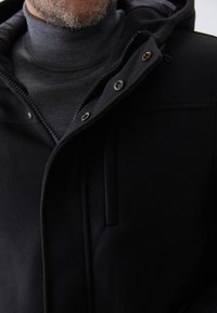 Chaqueta con capucha negra hecha de material texturizado, con cierre de cremallera, botones a presión y bolsillos laterales. Llevada sobre un suéter de cuello alto gris.