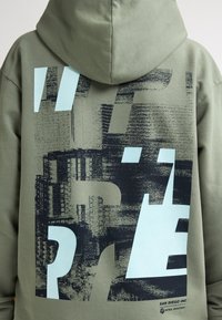 Olivgrüner Hoodie mit großen graphischen Drucken in Dunkelblau und Hellblau, mit strukturierten Mustern und Kängurutasche.