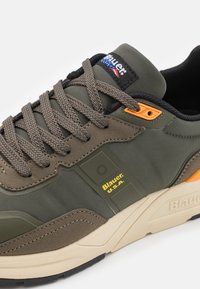 Zelená sportovní obuv s texturovaným síťovaným svrškem, béžovou gumovou podrážkou, šedými tkaničkami, oranžovými akcenty a černým interiérem. Viditelné logo "Blauer U.S.A.".