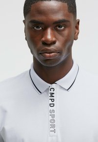 Polo shirt blanc avec un col à rayures noires et le texte "CMPD SPORT" sur le devant. Texture lisse, coupe classique.