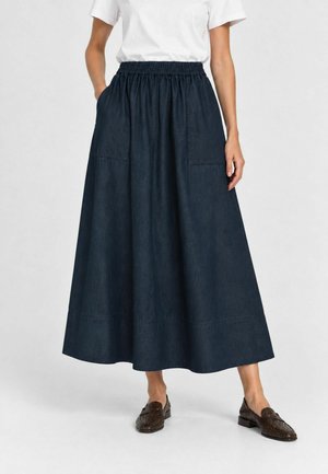 Persona con una falda midi azul oscuro de cintura alta y bolsillos delanteros, camiseta blanca y mocasines marrón oscuro, de pie contra un fondo claro.