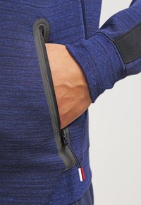 Giacca sportiva blu navy con finitura texturizzata, presenta una tasca laterale con cerniera nera e un dettaglio della linguetta di apertura rosso, bianco e blu.