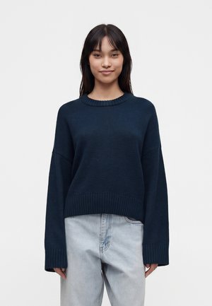 Junge Frau mit langen dunklen Haaren in einem dunkelblauen Pullover und hellblauen Jeans, die vor einem einfarbigen weißen Hintergrund steht.