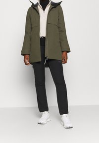 Olijfgroene waterdichte jack met een capuchon, twee voorzakken en een ritsluiting. Gecombineerd met zwarte broeken en witte sneakers.