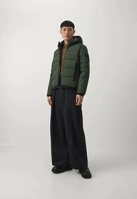 Mlada oseba stoji oblečena v zeleni pufasti jopič s kapuco, rjav srajco, široke črne hlače in črne čevlje pred enobarvno belo ozadje.