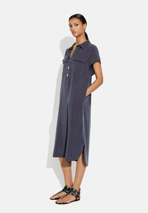 Kobieta stoi z rękami w kieszeniach, ubrana w ciemnoszarą sukienkę midi z krótkim rękawem, z guzikami z przodu i czarne sandały z ćwiekami.